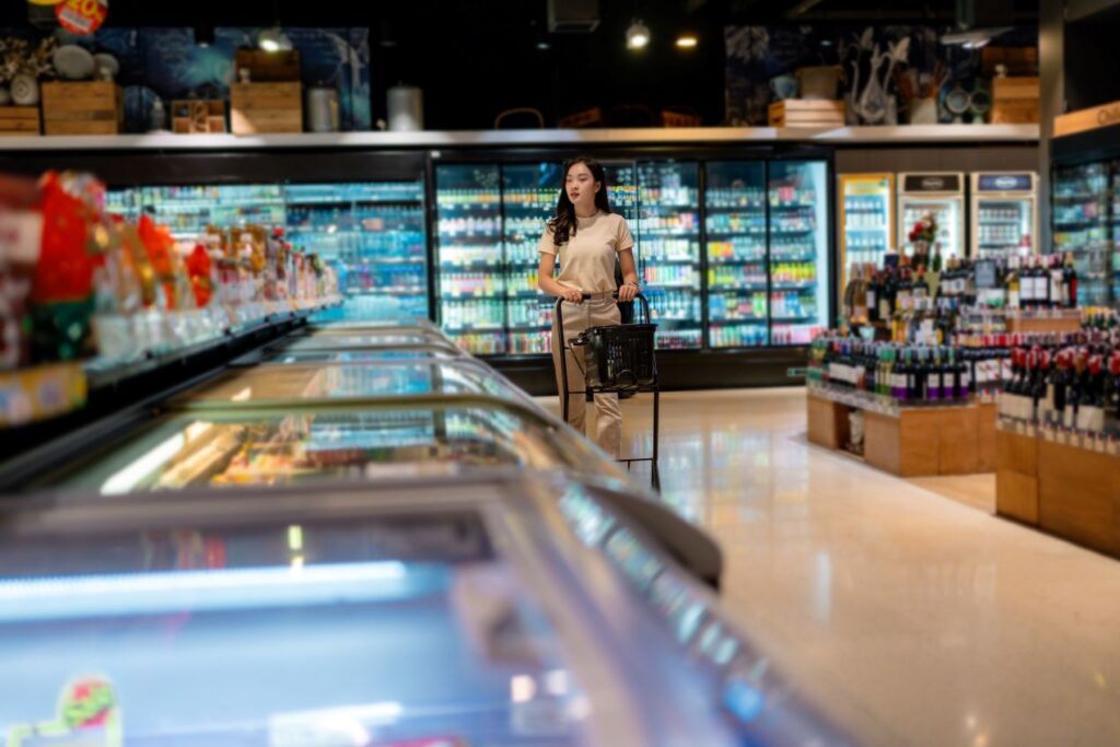 Kundin mit Einkaufskorb geht an moderner Kuehlvitrine im Supermarkt entlang – Frischezone im Fokus der Raumgestaltung